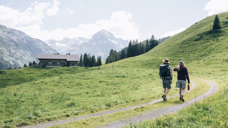 Zwei Wanderer auf einem gewundenen Weg in einer grünen Landschaft mit Bergen im Hintergrund