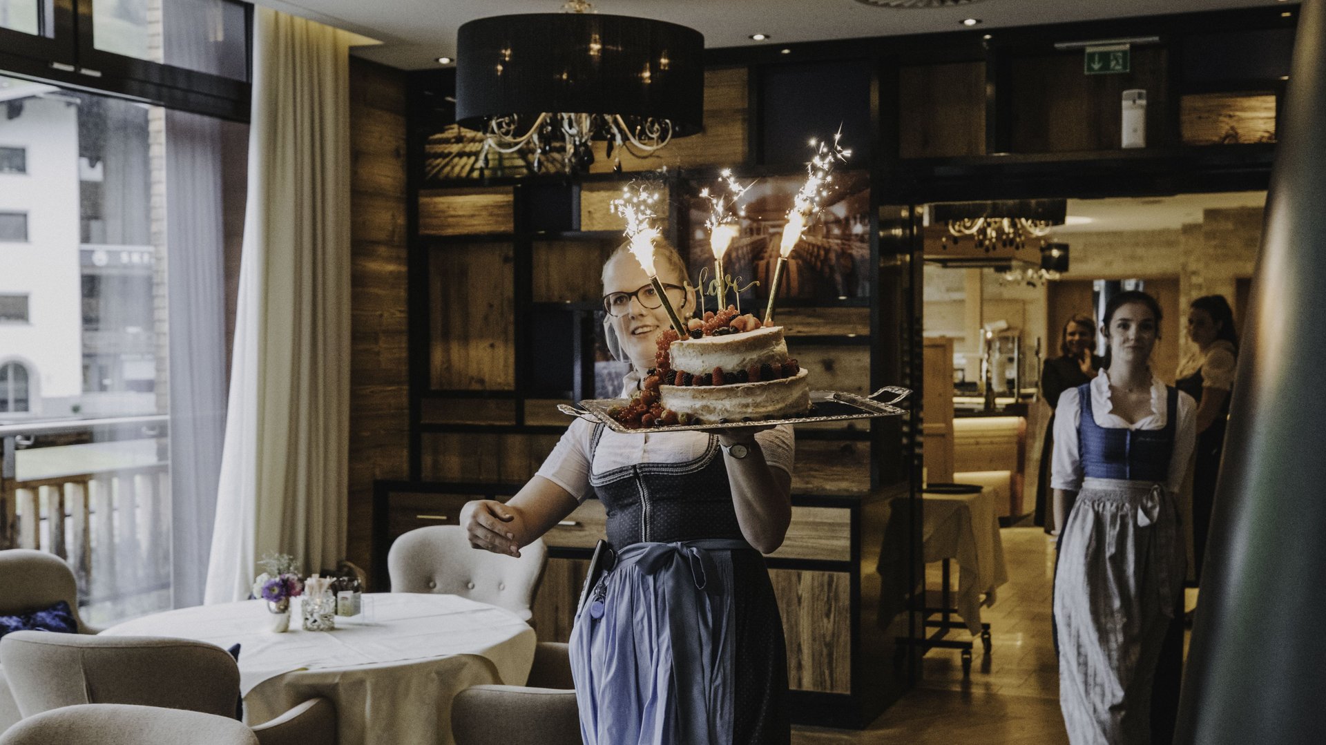 Frau im Dirndl trägt Kuchen mit Wunderkerzen in elegantem Restaurant.