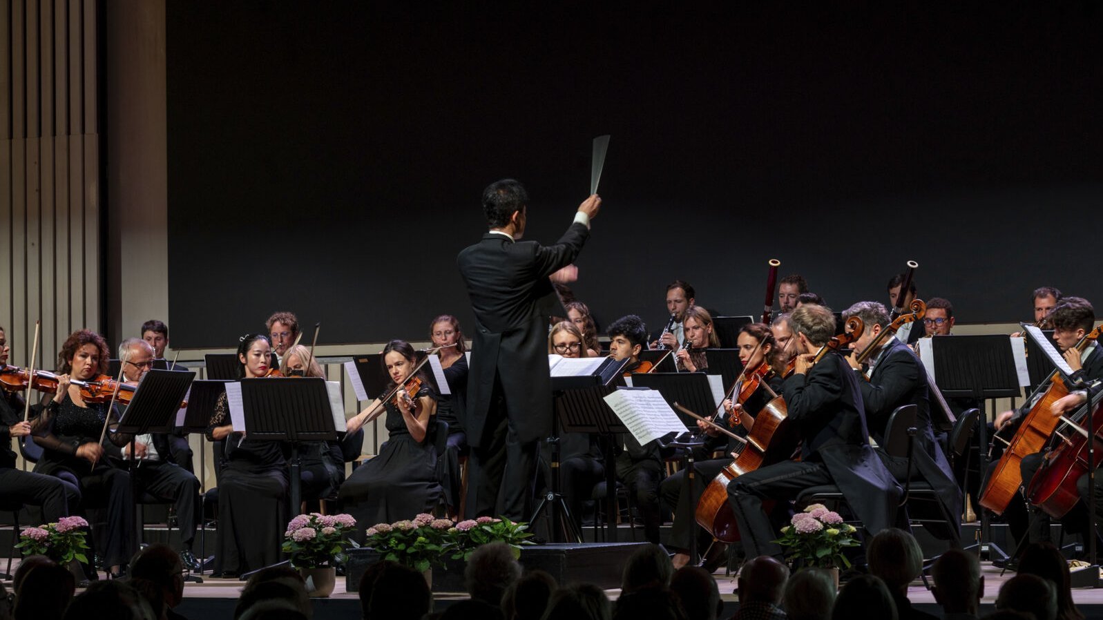 Orchester mit Dirigent bei einer Aufführung auf der Bühne, Publikum im Vordergrund sichtbar