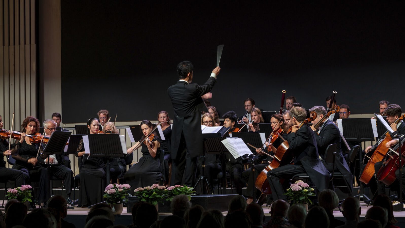 Orchester mit Dirigent bei einer Aufführung auf der Bühne, Publikum im Vordergrund sichtbar