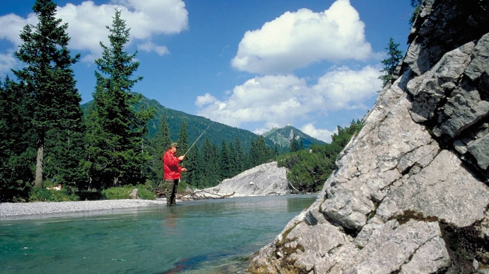 Person fishing in a clear mountain river, surrounded by trees and rocks under a blue sky.