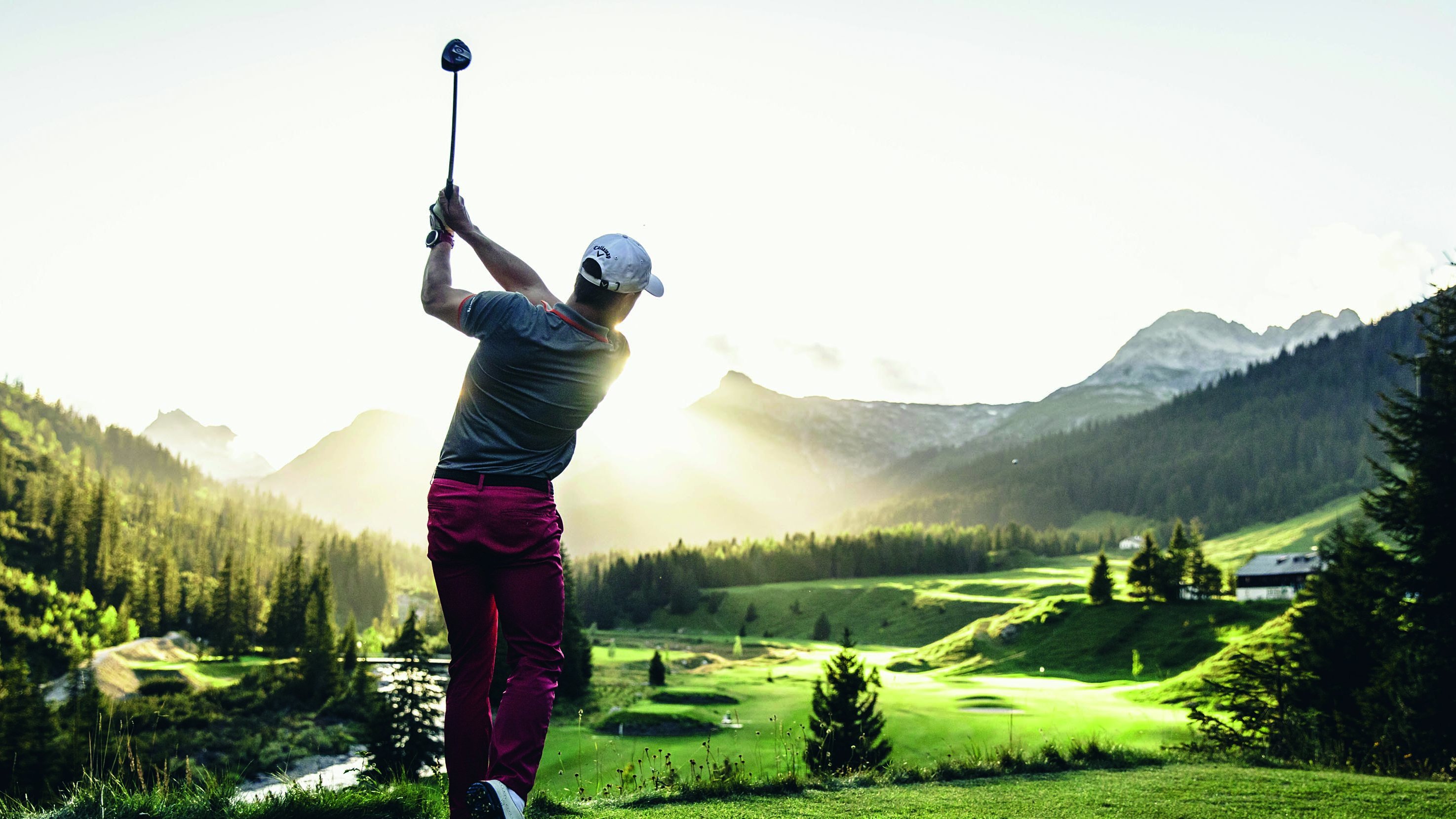 Golfer schwingt den Schläger bei Sonnenuntergang in einer beeindruckenden Berglandschaft.