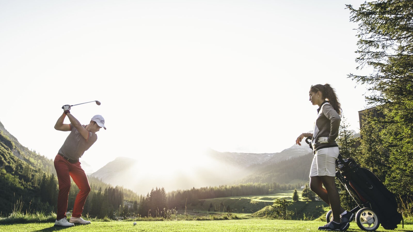 A person takes a swing on a golf course with mountains in the background while a woman observes nearby.