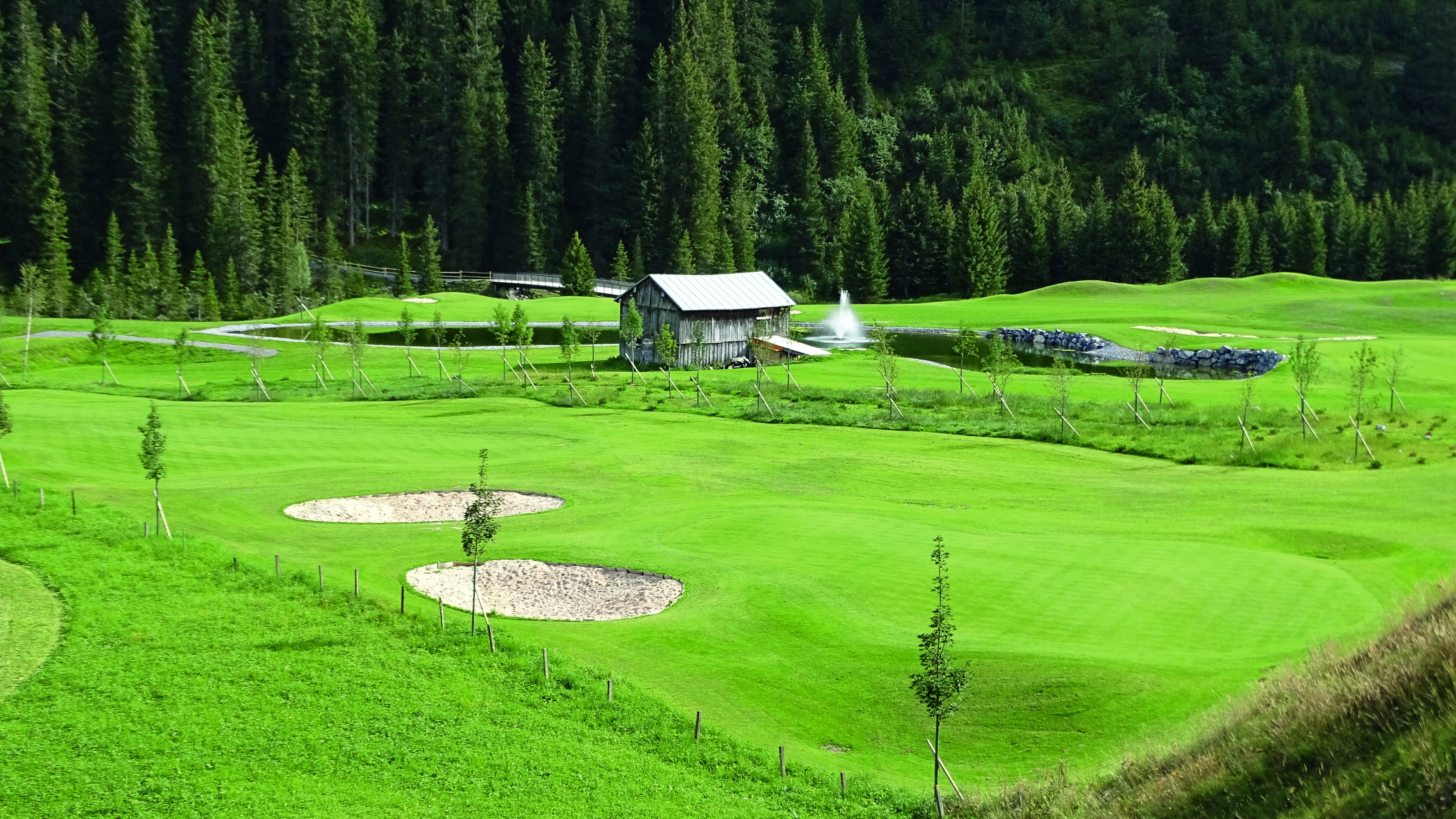 Grüner Golfplatz mit rustikalem Gebäude und Wald im Hintergrund.