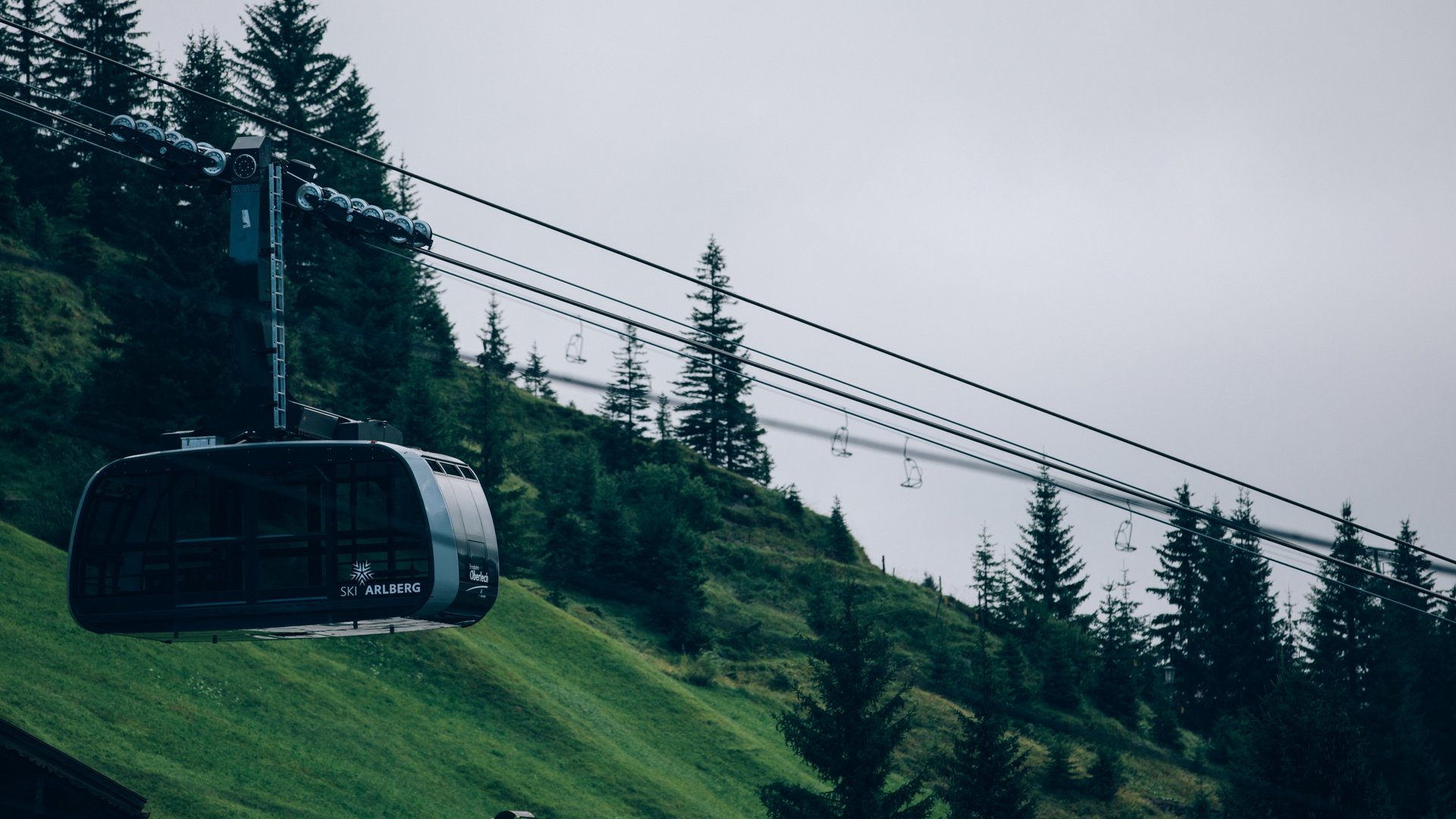 Cable car floats over green slopes in front of wooded hills and cloudy skies.
