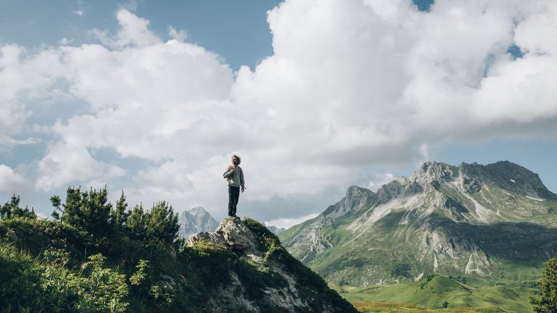 Eine Person steht auf einem felsigen Vorsprung in einer Berglandschaft, umgeben von Bäumen und Wolken.