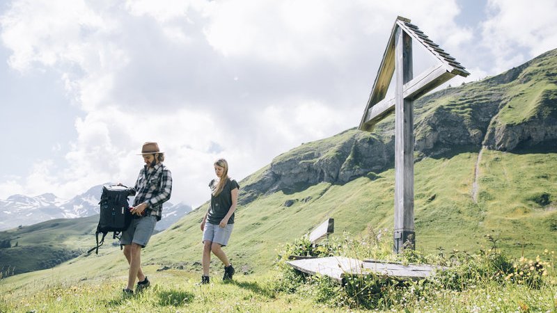 Zwei Wanderer auf einem Pfad mit einem Holzkreuz in der Vordergrund und grünen Hügeln im Hintergrund.