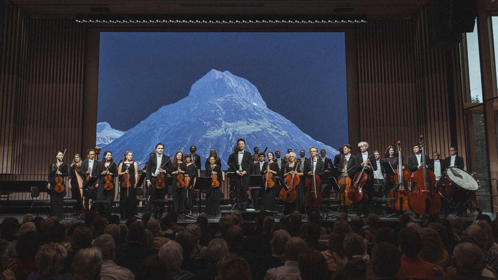 Orchestra Performance Against Majestic Mountains An orchestra plays on stage with a large mountain backdrop behind them and an audience in front.