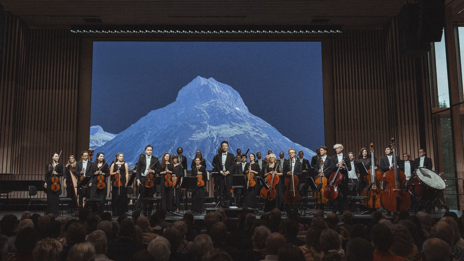 Orchesterperformance vor einer großen Bergkulisse mit Publikum im Vordergrund und Musikern auf der Bühne.