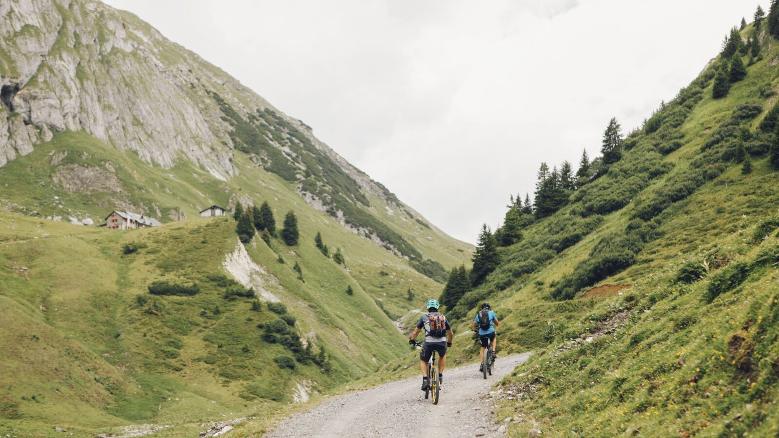 Two cyclists navigating a dirt path in a mountainous landscape with green hills and rocky cliffs