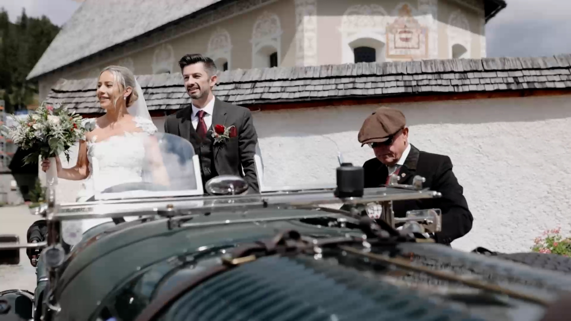 Braut und Bräutigam sitzen in einem Oldtimer, während ein Fahrer in einer Mütze vorne sitzt. Rustikale Gebäude im Hintergrund